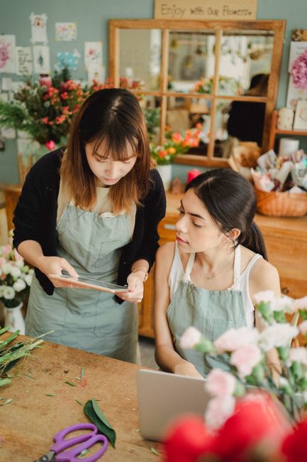 In a flower shop, two women are engaged in a floral arrangement activity. The woman standing has shoulder-length dark hair with light brown highlights and is wearing a black cardigan over a light blue