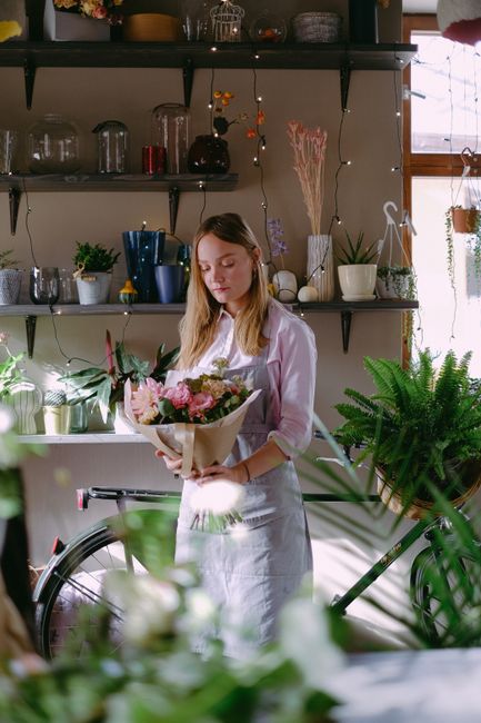 A young woman with long light brown hair, dressed in a white shirt and apron, is holding a bouquet of mixed flowers wrapped in light brown paper. The bouquet features pink roses, purple orchids, and g
