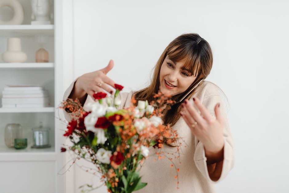 A woman smiling and holding a vibrant floral bouquet consisting of various fresh flowers such as red roses, white lisianthus, and orange hypericum berries, with green foliage. She appears to be engage
