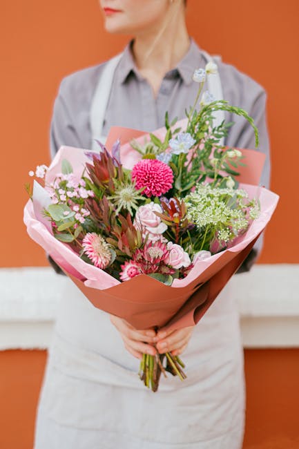 A person dressed in a light-colored shirt and white apron holding a large bouquet of fresh flowers wrapped in pink and peach tissue paper. The bouquet contains a mix of pink, white, and purple blooms,