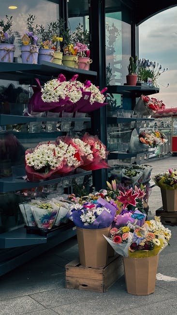 A display of fresh floral arrangements on a multi-tiered black metal shelving unit outside Eastham Flowers, situated on Station Road CH62. The arrangements include vibrant bouquets wrapped in pink, pu