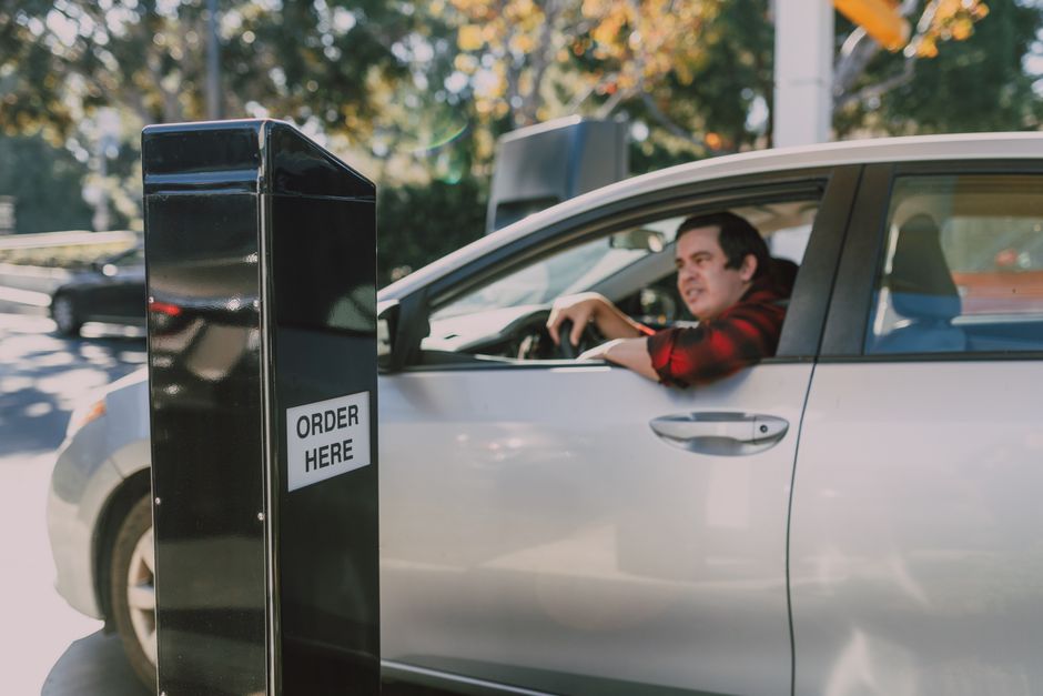 A man is sitting in a silver car with his arm resting on the window as he prepares to receive a floral gift from a black flower delivery box marked 'ORDER HERE' in an outdoor setting with trees and ot