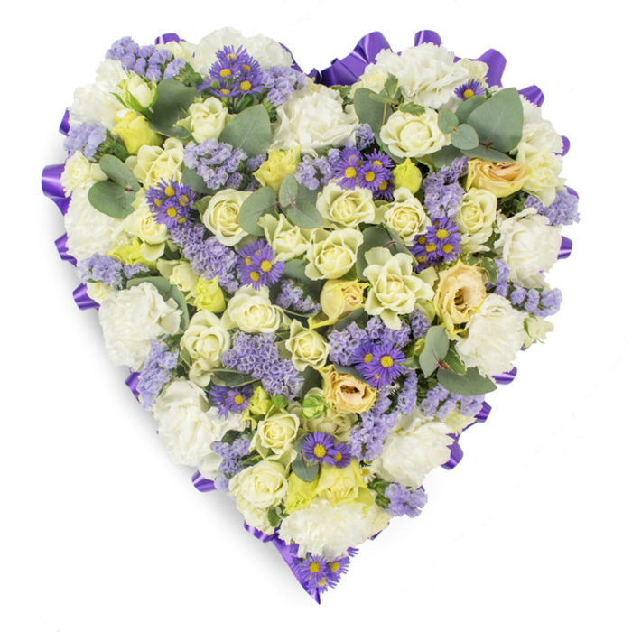 Set against a stark white background in a studio-style image, this heart-shaped condolence arrangement is a study in gentle contrasts and expert floristry. The entire perimeter is traced with a continuous band of vivid purple satin ribbon, neatly looped to define the heart's shape and lend a formal, respectful edge to the design. Filling the interior is a dense, harmonious mix of soft whites, creams and pale yellows: numerous cream-to-pale-yellow roses, from compact buds to softly open blossoms, form the main body and give the piece a plush, tactile quality. Complementing them, clusters of white carnations add classic volume and a soothing, familiar floral texture. Sprays of lavender statice bring delicate mauve accents that lift the palette, while small purple aster-like blooms with prominent yellow centres create bright focal points and add visual depth across the heart. Interspersed throughout, rounded eucalyptus leaves in blue-green tones provide cooling contrast and a sculptural, leafy rhythm that breaks up the floral mass. The arrangement reads as an elegant funeral tribute - calm, composed, and designed to convey remembrance and respect at a church service, chapel, or graveside in East Ham or nearby Manor Park. Its serene colourway and careful construction reflect the hands of an experienced florist who has balanced texture, scent and form to offer a dignified expression of sympathy and enduring love.