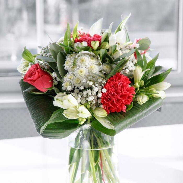A refined red and white bouquet is captured here in a moment of fresh, just-arranged beauty, standing tall in a clear cylindrical glass vase whose transparency reveals a cluster of neatly cut green stems resting in clean water. The design is anchored by velvety deep red roses and a standout crimson carnation, the carnation's densely ruffled petals forming a rich, textured centrepiece that draws the eye forward. Around these passionate red tones, a soft halo of white and cream blooms creates harmony: slender-petalled white alstroemeria flecked with fine markings, rounded chrysanthemum-style flowers, and several poised white lily buds still closed, promising a slow, elegant unfolding over the days ahead. Delicate sprigs of gypsophila, or baby's breath, thread throughout the composition, their tiny white clusters adding an ethereal, cloud-like lightness that softens the stronger shapes. The bouquet is held together and visually grounded by generous greenery. Broad, glossy dark green leaves are carefully curled and wrapped at the base, forming a tailored collar that feels both modern and classic, while taller, eucalyptus-like stems and pointed foliage lift the eye upward and add nuanced shades of green from deep forest to cool grey-green. The arrangement is set on a smooth white surface, with a bright, softly blurred background that suggests daylight streaming through a window-perhaps in an East Ham townhouse near Plashet Park-casting gentle highlights on each petal and leaf. The mood conveyed is thoughtful and uplifting, a balance of romance and serenity that would make this bouquet a graceful centrepiece for a family celebration, an anniversary dinner, or a heartfelt gesture of gratitude delivered locally by East Ham Flowers to homes and offices across the neighbourhood.